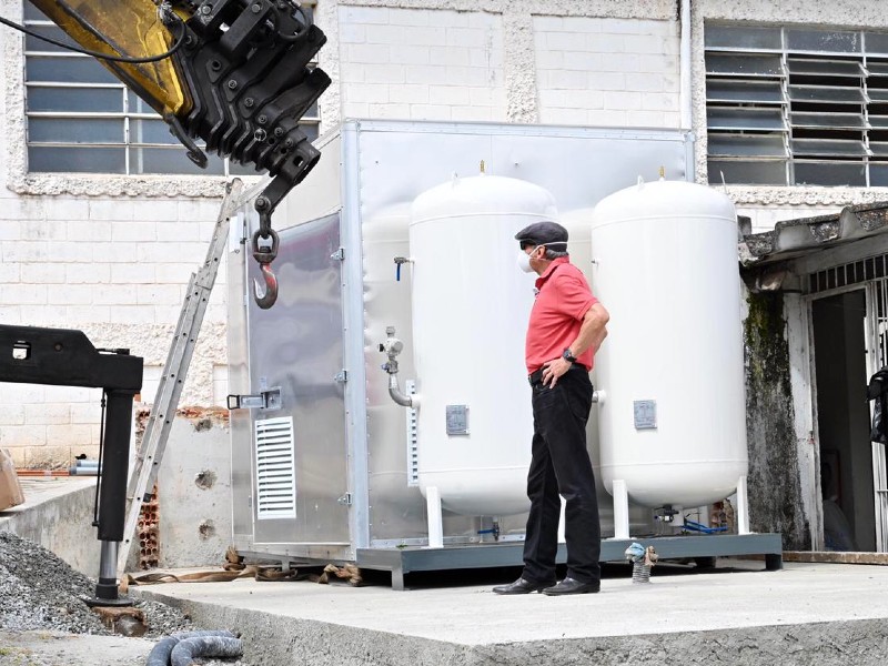 Foto da miniusina de oxigênio no HM Capela do Socorro. Ela é composta por um container metálico e dois cilindros grandes, brancos, na parte de fora do container.